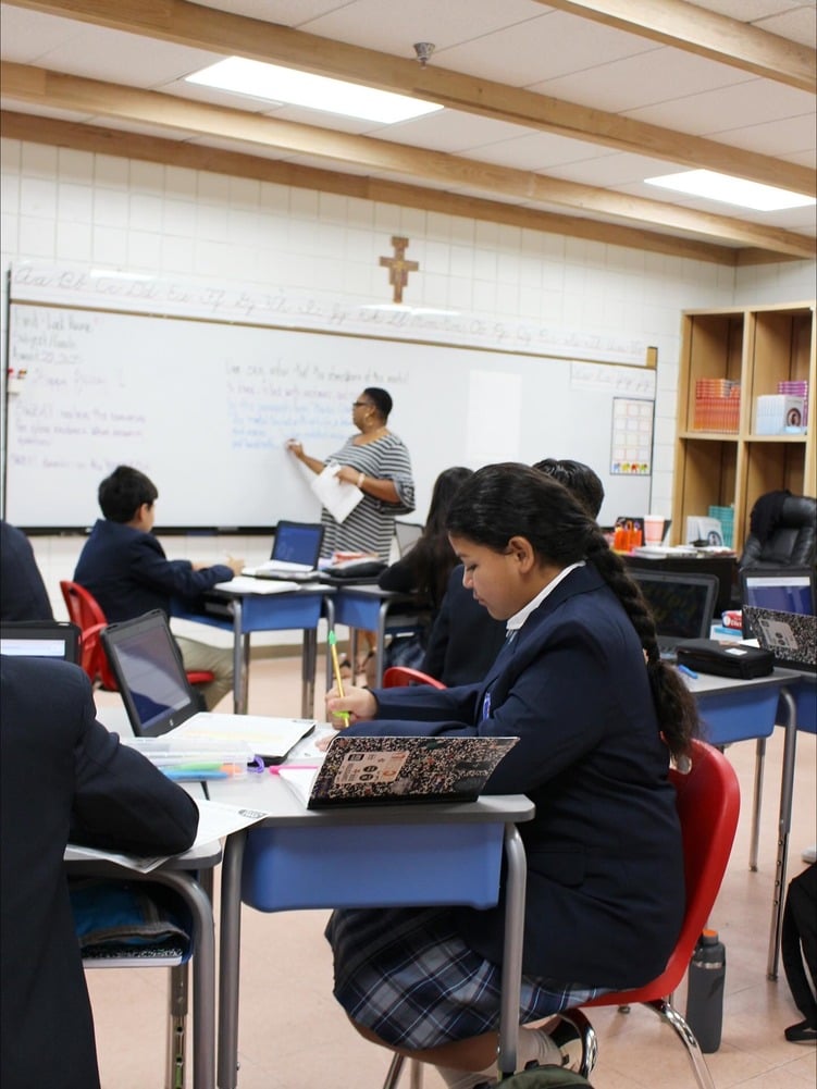 Students in uniform work at desks while teacher writes on whiteboard