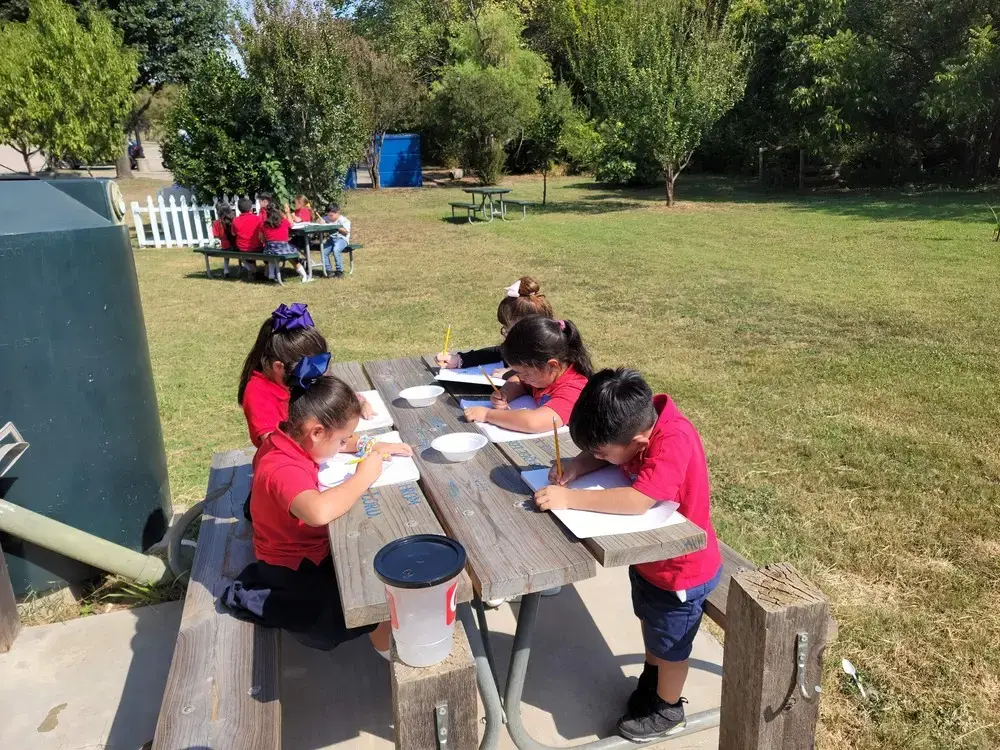 Students in red uniforms writing at outdoor picnic tables