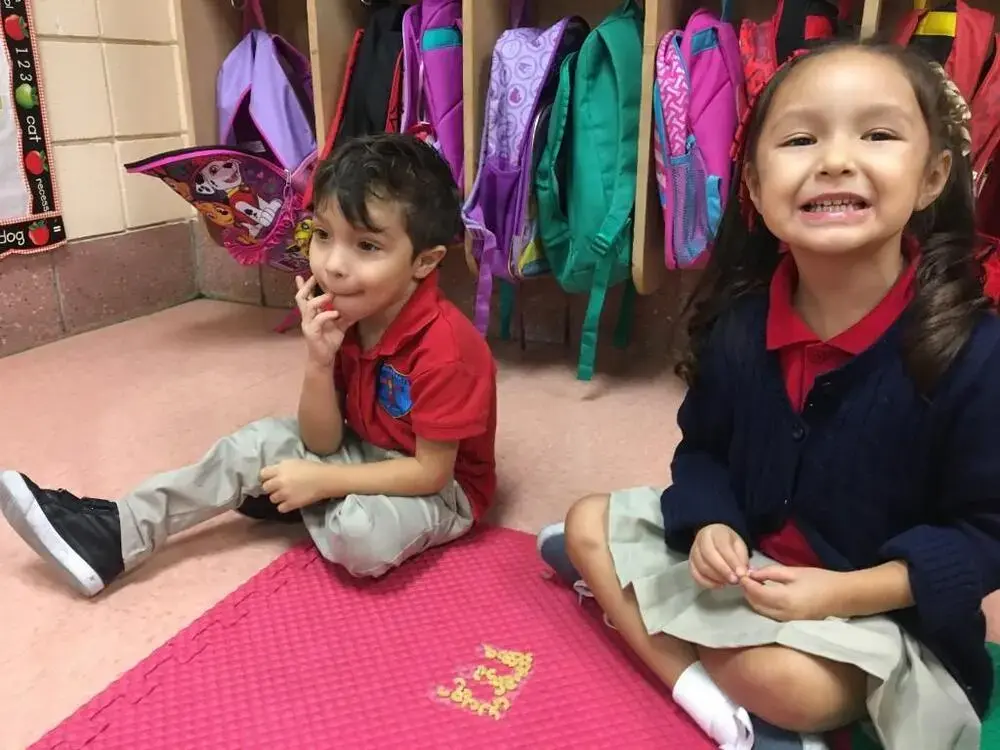 Two young students in uniforms sitting on classroom floor smiling