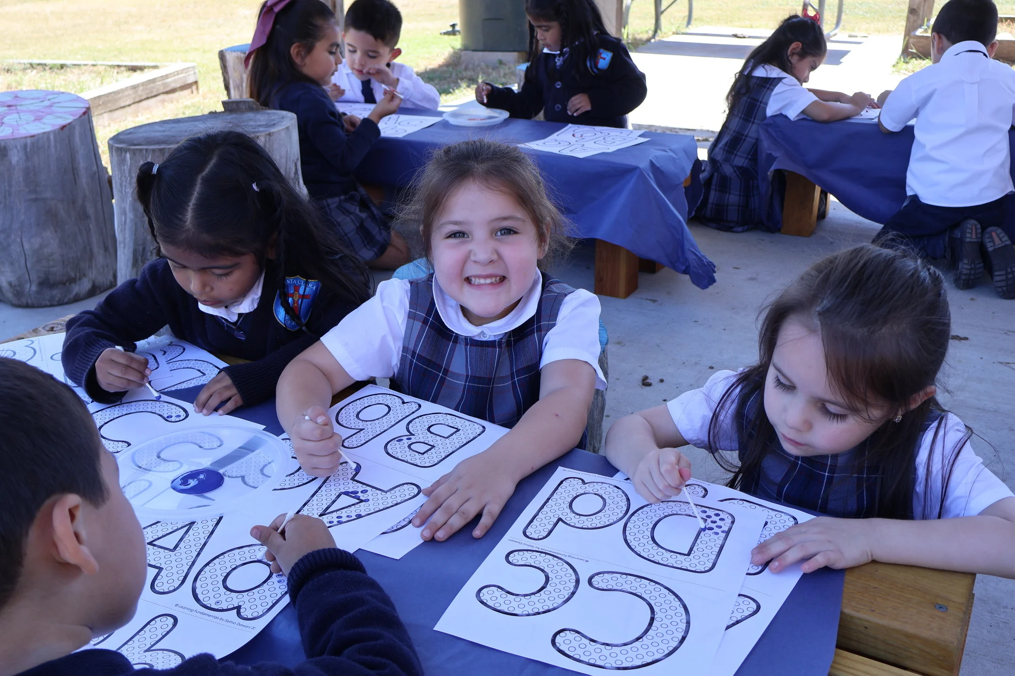 Students in school uniforms completing alphabet letter worksheets outdoors