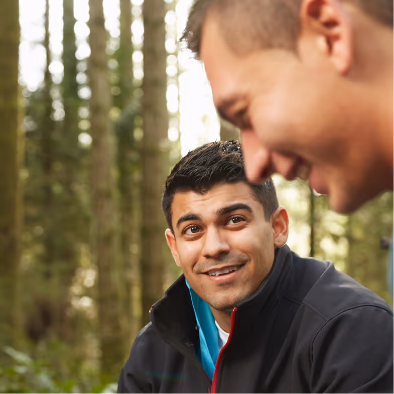 Two men outdoors in a forest, one smiling and looking up at the other.