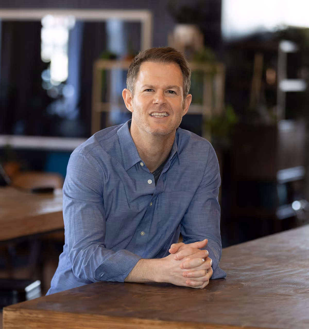 Smiling man in a blue button-up shirt sitting at a wooden table in a cozy indoor setting.