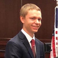 Young man in a suit and red tie standing near an American flag indoors.