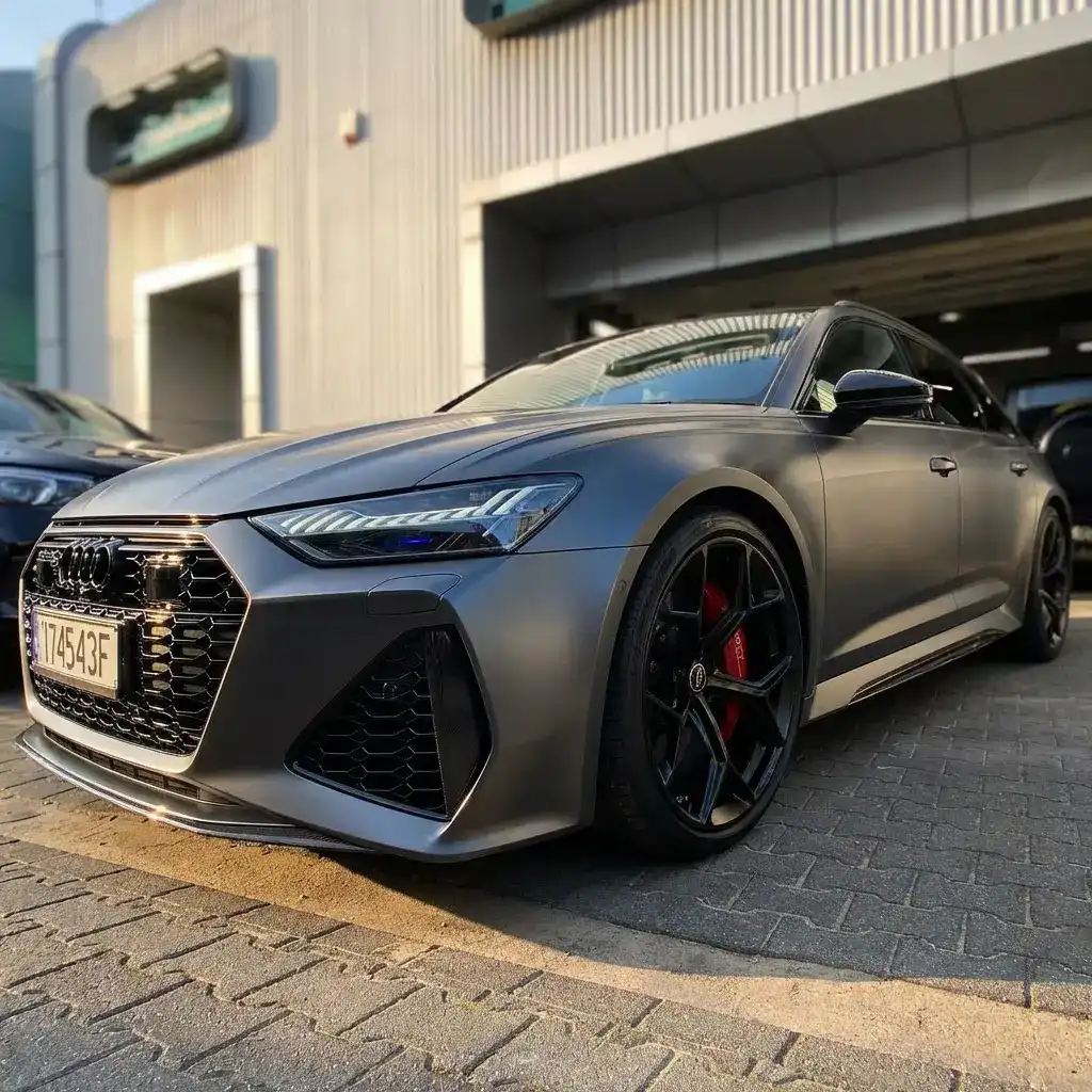 Audi RS6 w kolorze grafitowym, oklejona przezroczystą folią matową PPF, widok od przodu i z boku, na tle salonu samochodowego.
