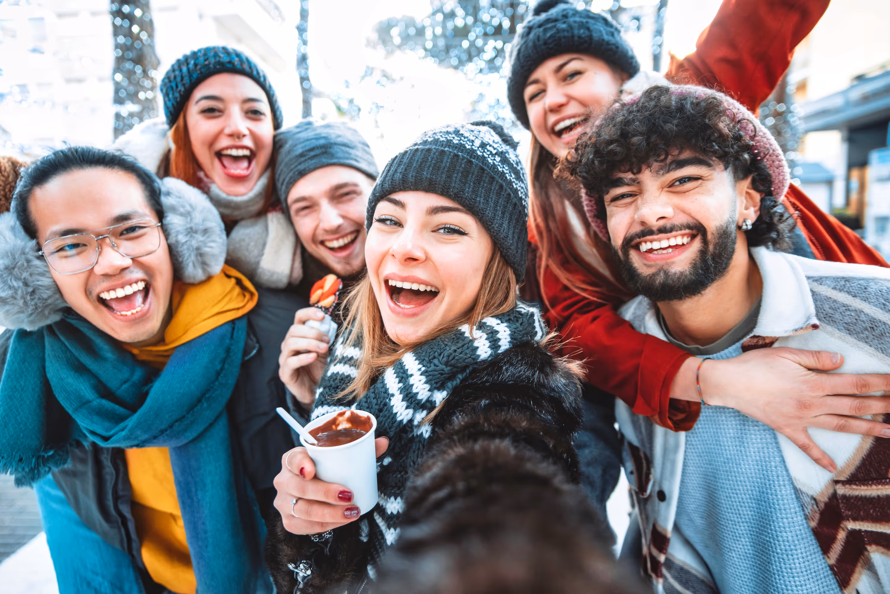A group of smiling residents posing for a selfie in an outdoor urban setting decorated with festive holiday lights, holding warm beverages near The Eleanor Daytona Beach.