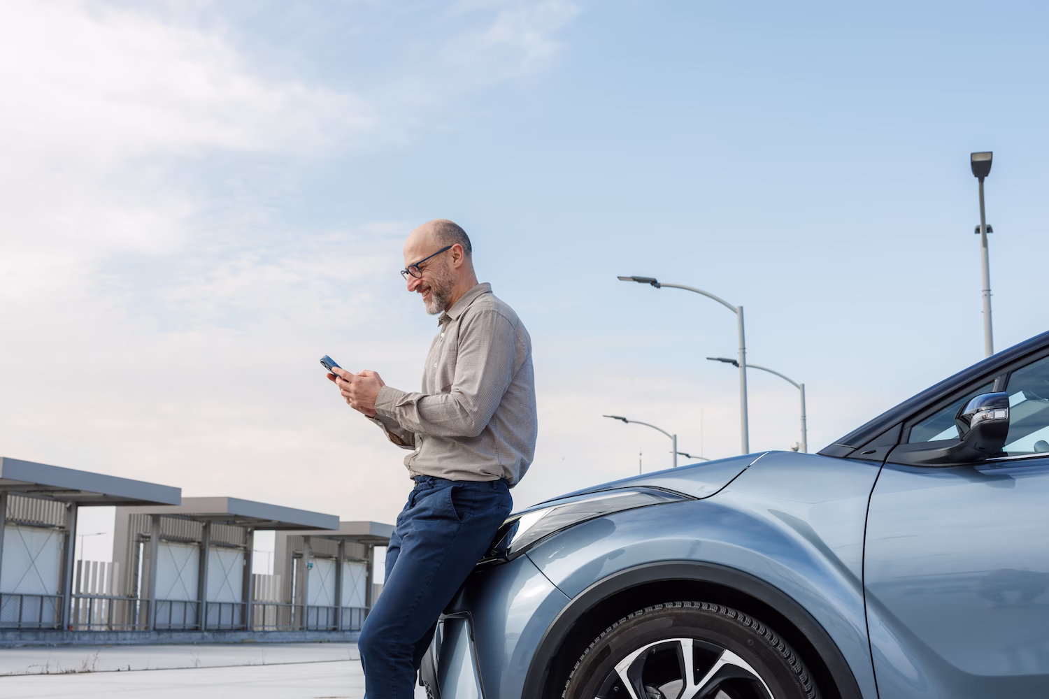 Smiling man with glasses leaning on a blue car while using a smartphone outdoors under a clear sky.