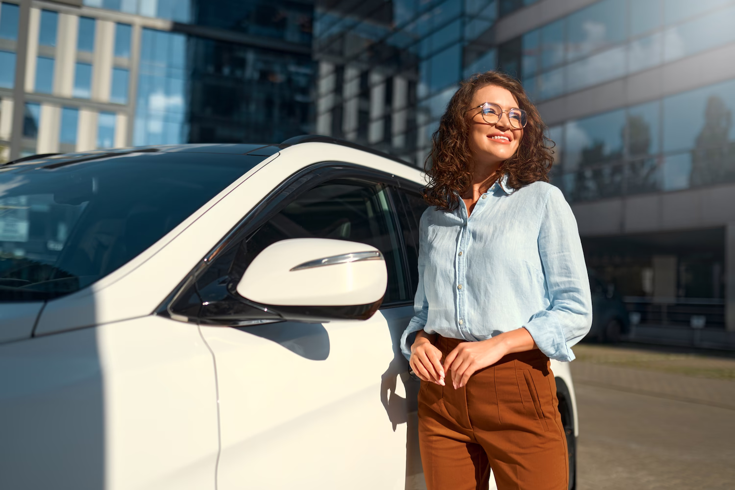 Smiling woman with curly hair wearing glasses, a light blue shirt, and brown pants leaning against a white car outside a modern glass building.