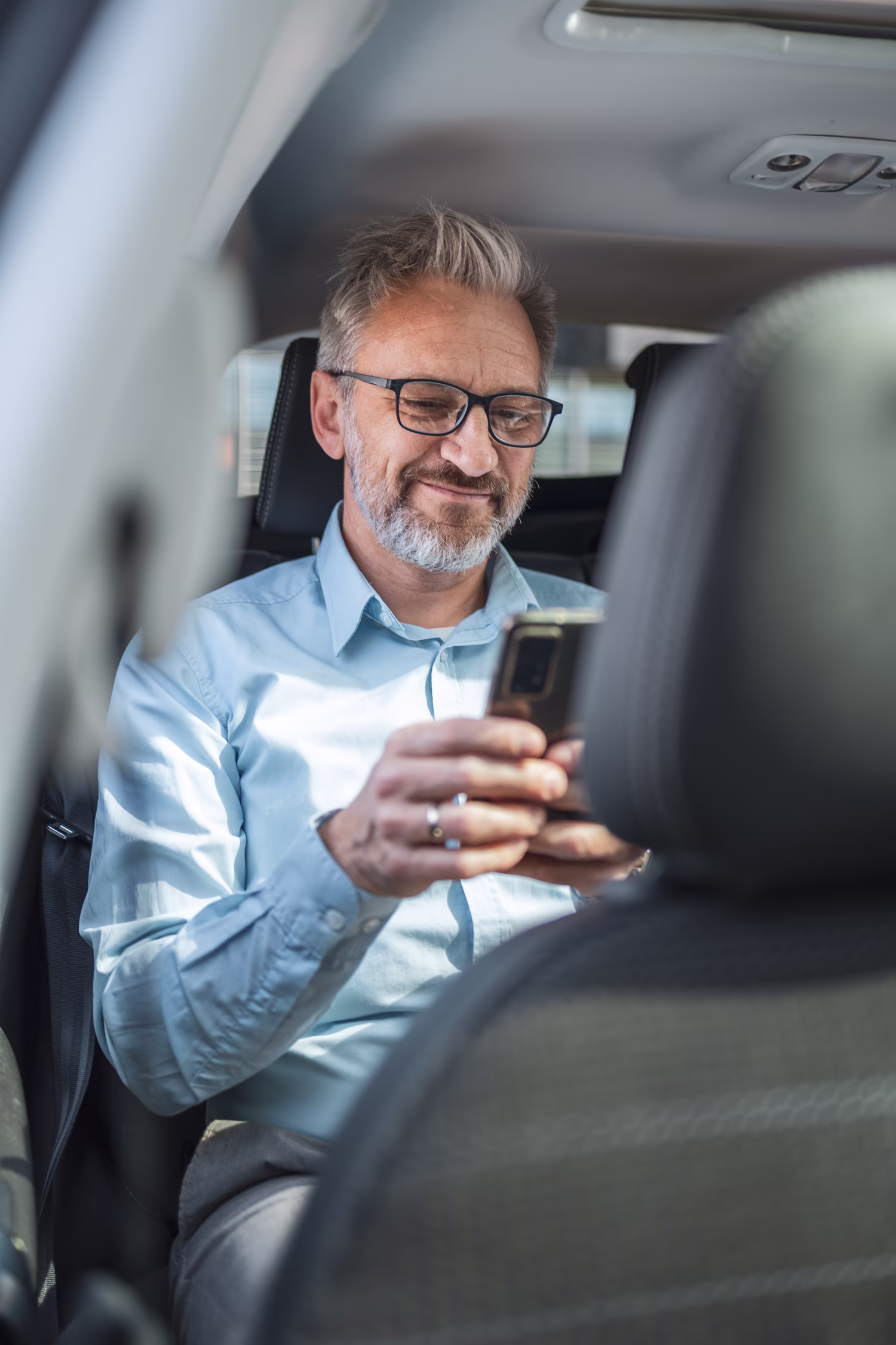 Middle-aged man with glasses sitting in the backseat of a car looking at his smartphone and smiling.