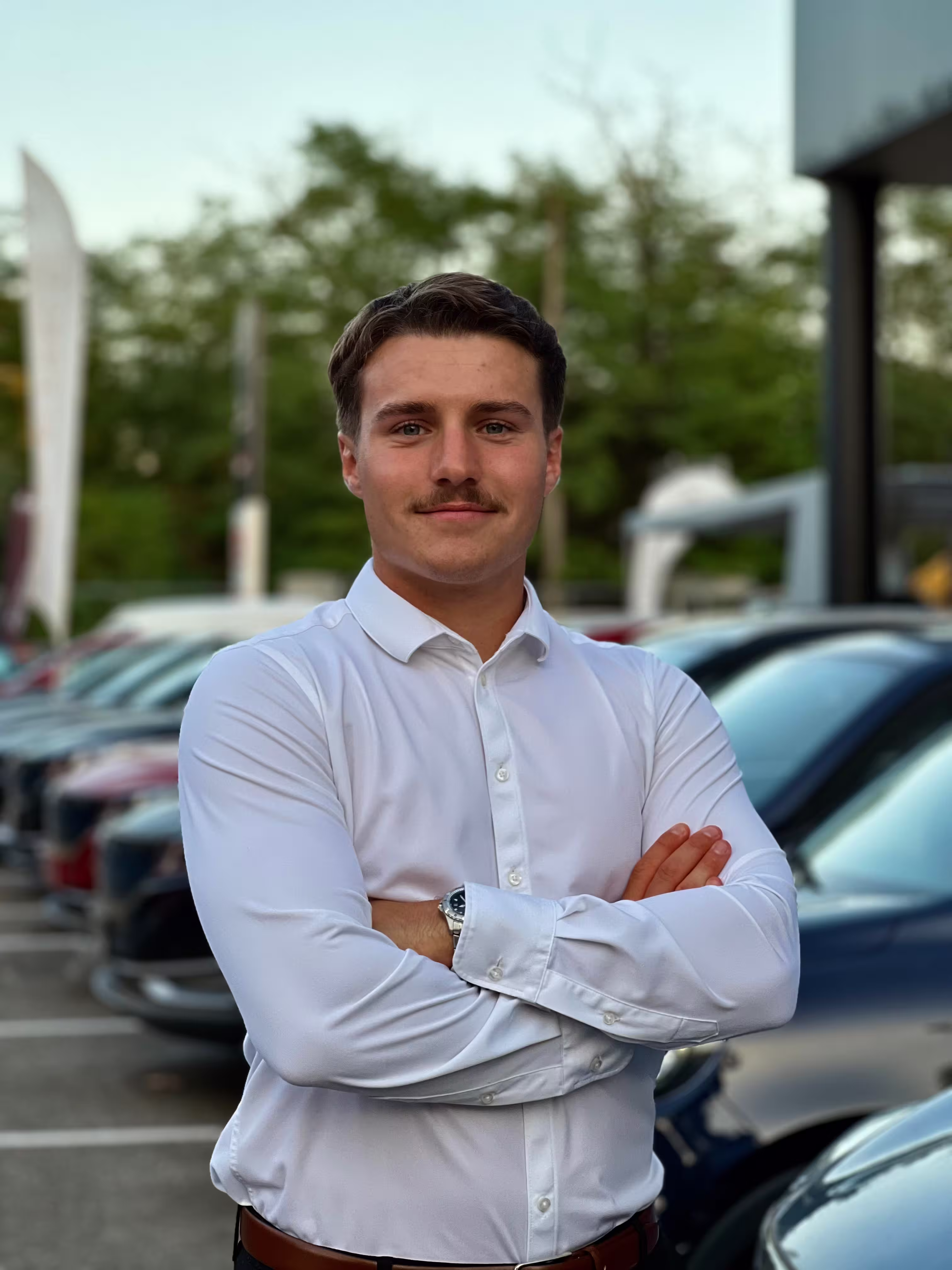 Jeune homme avec chemise blanche et montre, bras croisés, debout devant une rangée de voitures en extérieur.