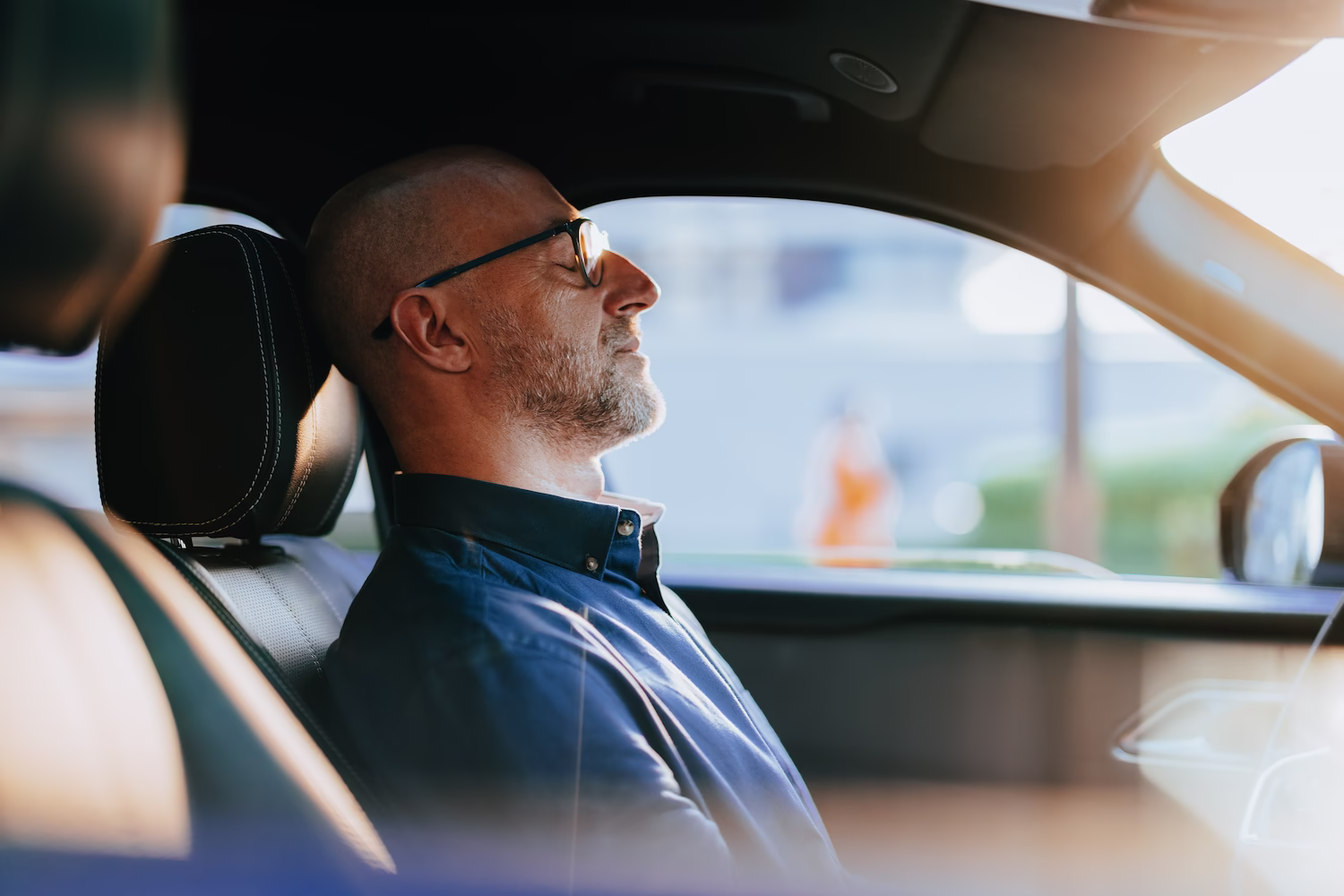 Man with glasses sitting peacefully in the driver's seat of a car with eyes closed.