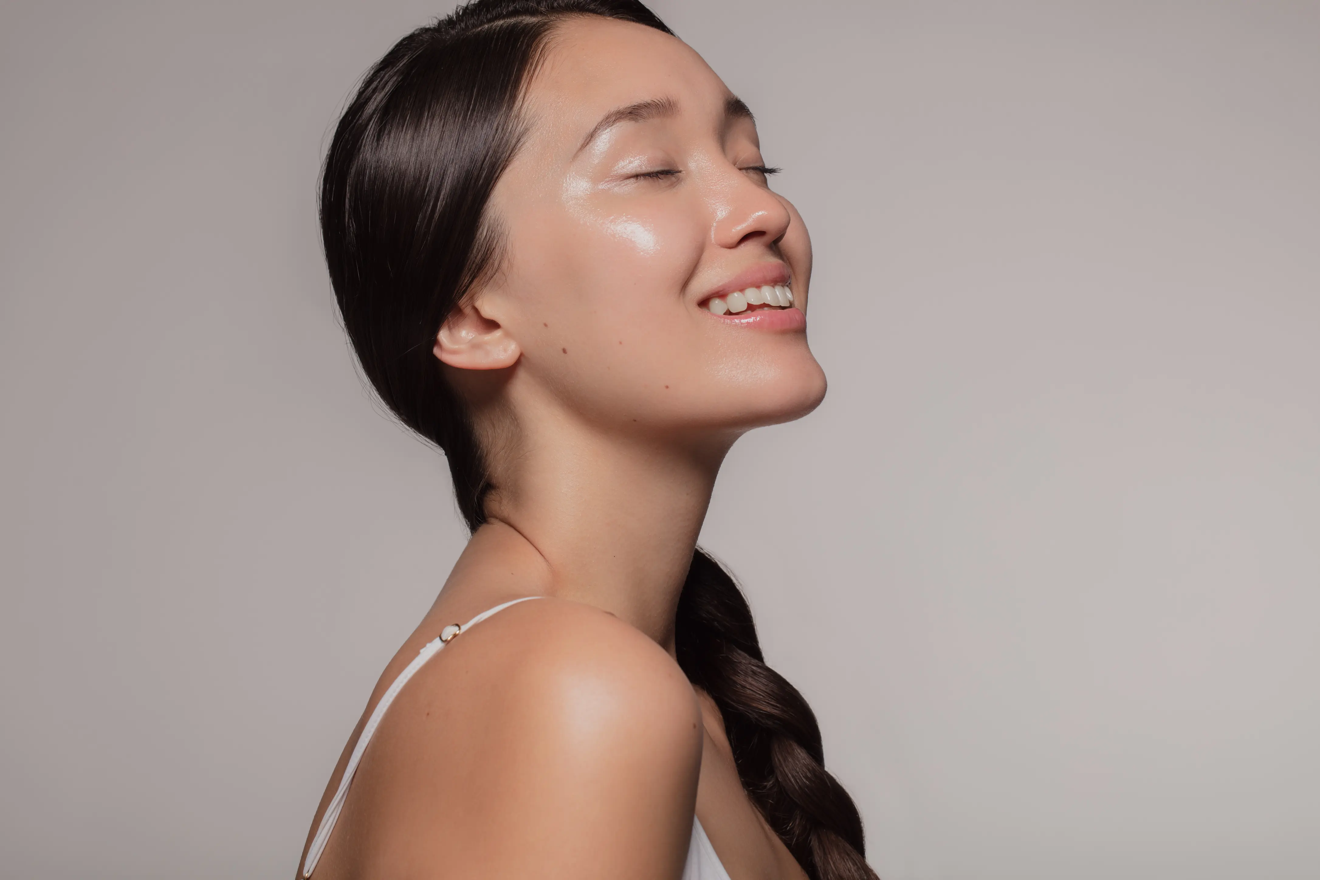 Smiling woman with closed eyes, smooth skin, and long braided hair wearing a white strap top.