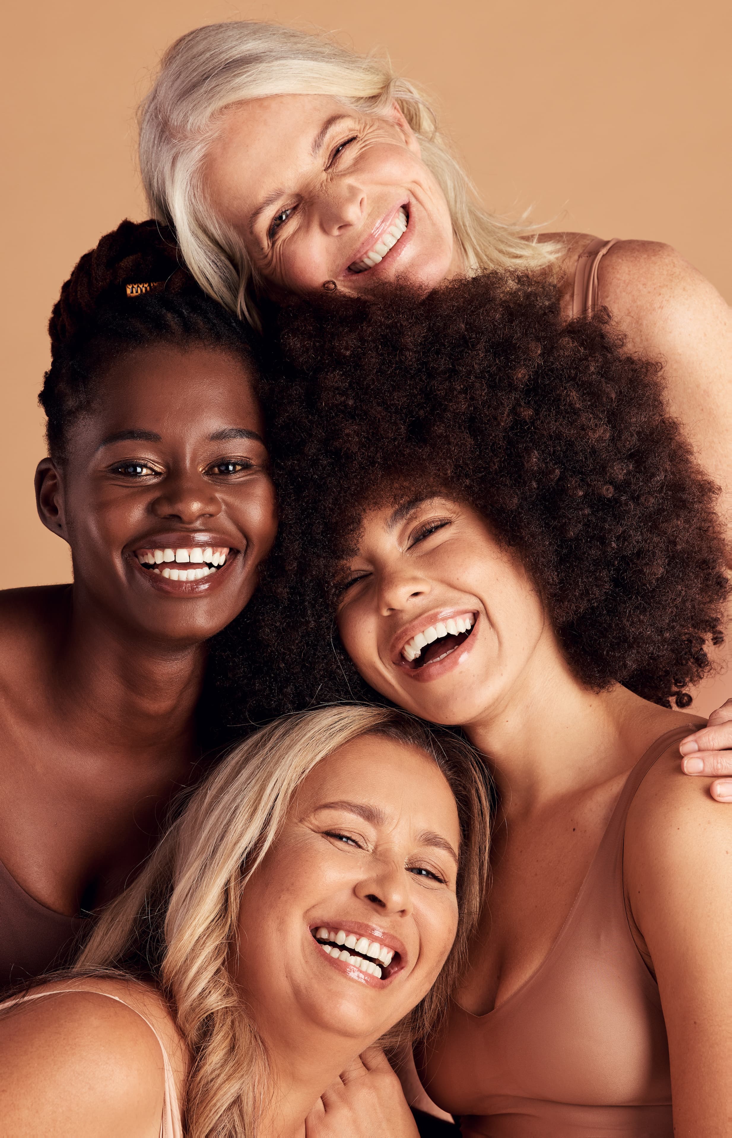 Four diverse smiling women in skin-toned tops close together against beige background.