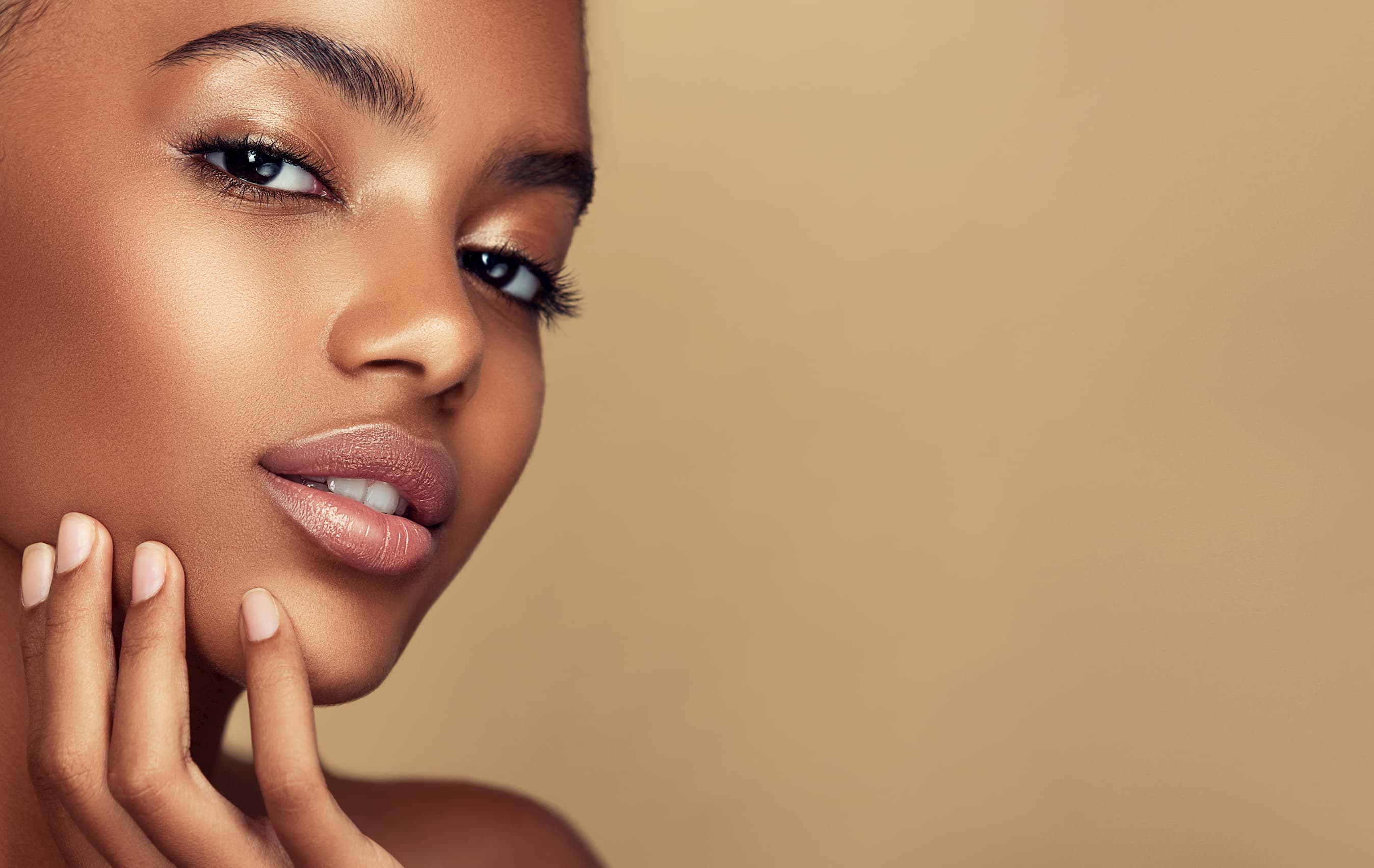 Close-up of a woman with smooth skin touching her face against a beige background.