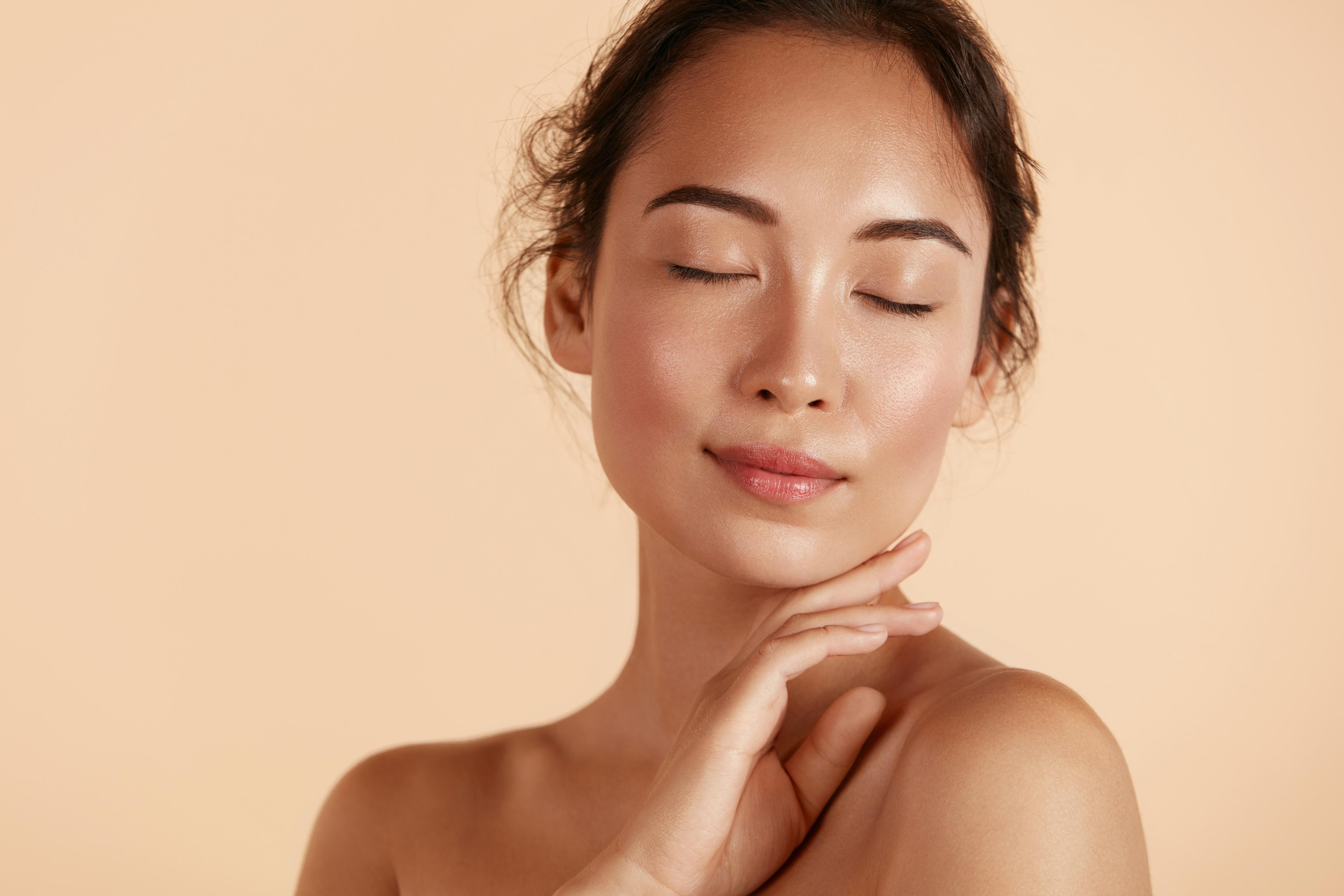 Young woman with clear skin and closed eyes gently touching her neck against a beige background.