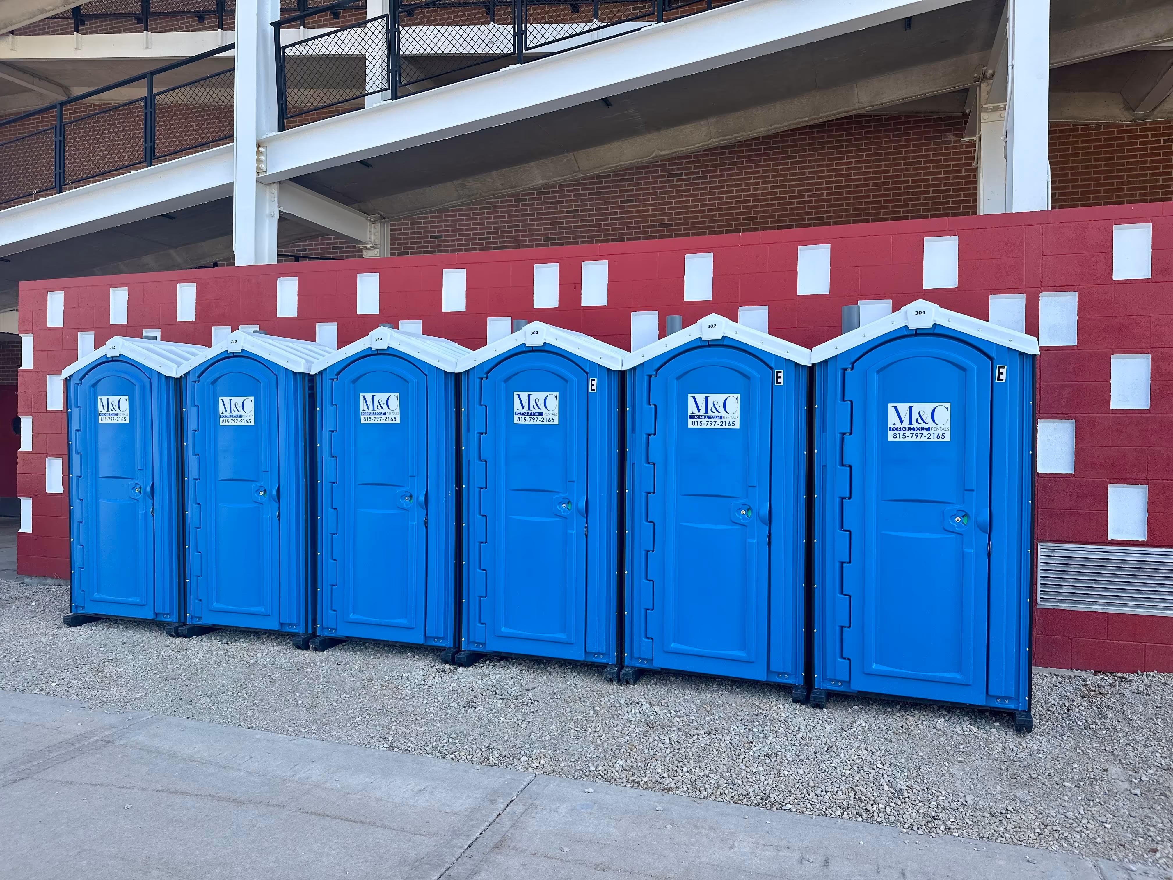 Six blue M&C portable toilets lined up against a red and white brick wall under a metal staircase.