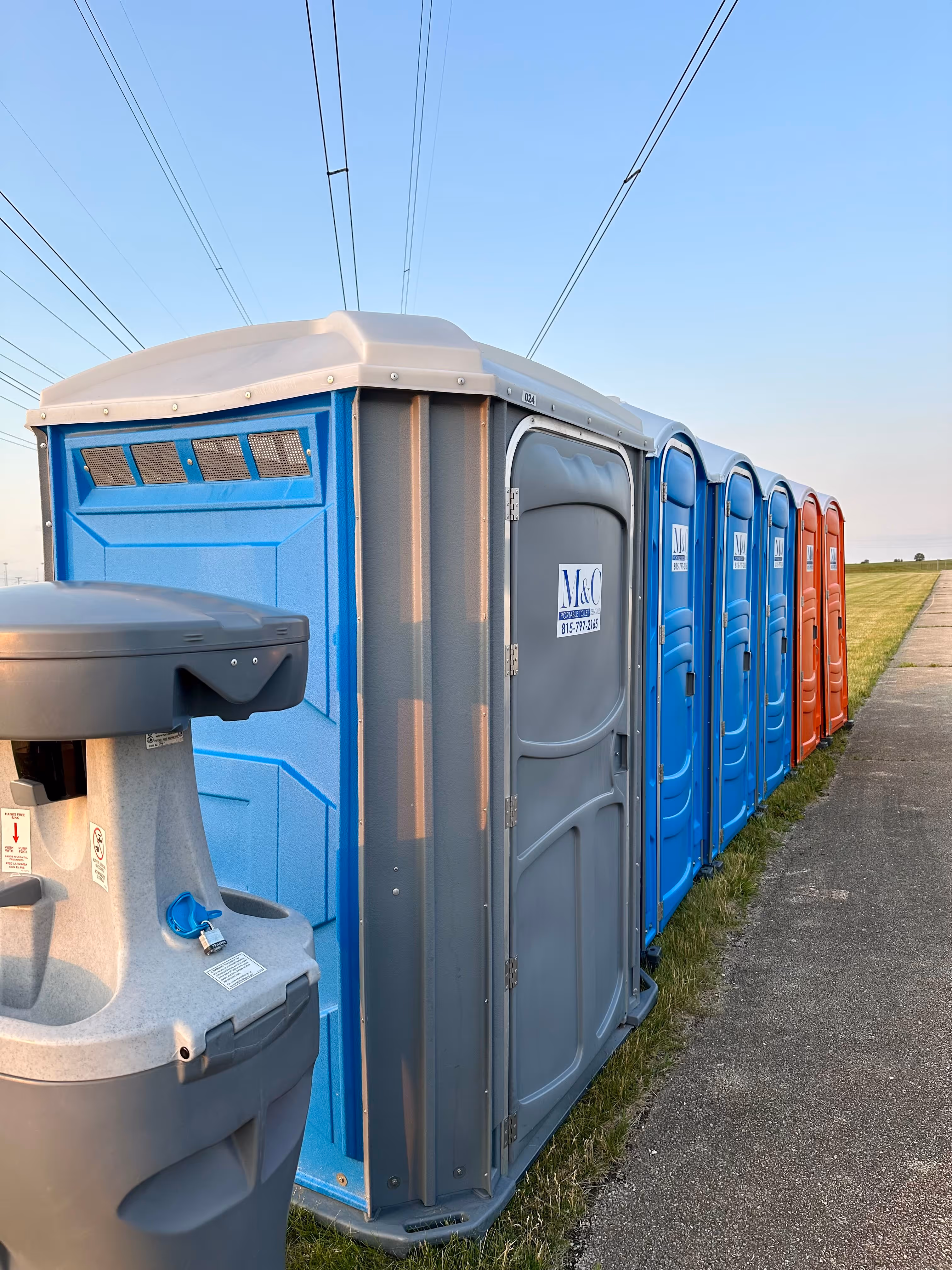 Row of portable toilets in blue, gray, and red colors lined up beside a paved path with a portable handwashing station in the foreground.