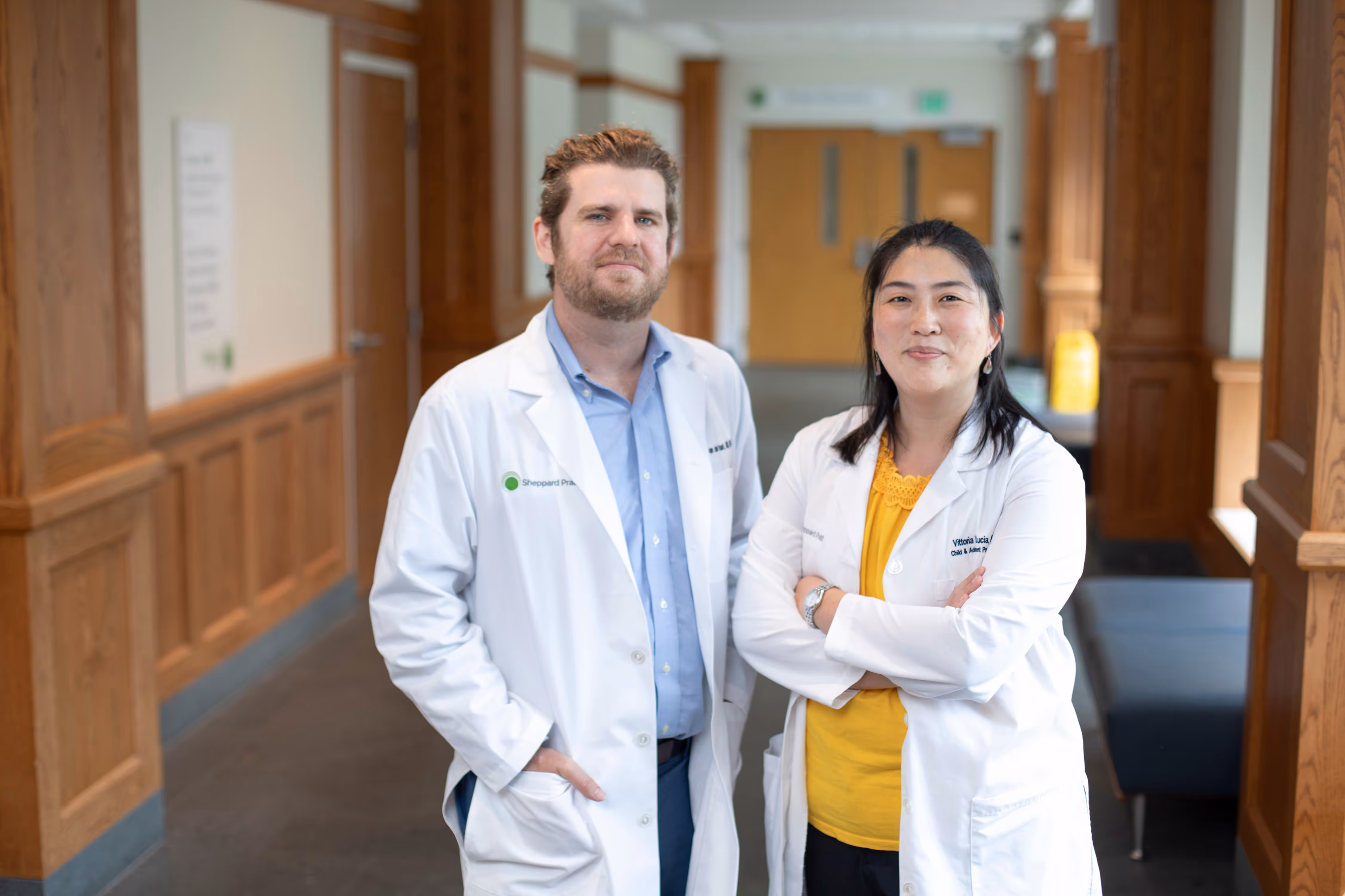 Two doctors in white coats standing in a hospital hallway, one with arms crossed and one with hands in pockets.