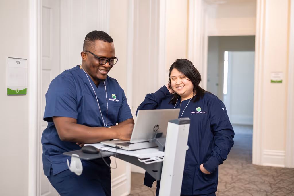 Two healthcare workers in navy scrubs smiling while looking at a laptop on a mobile cart in a hallway.