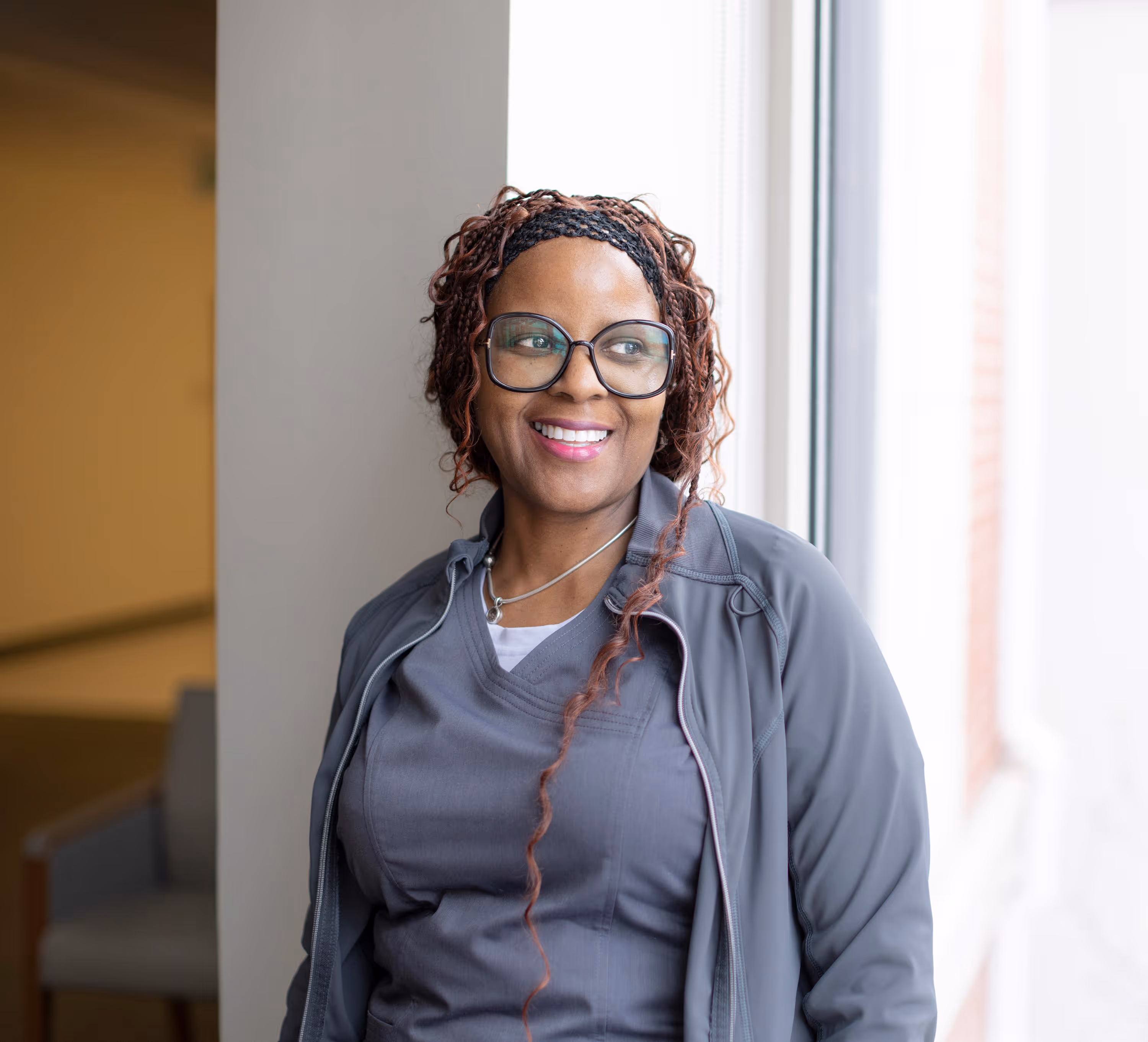 Smiling woman with glasses wearing gray scrubs and jacket standing by a bright window.
