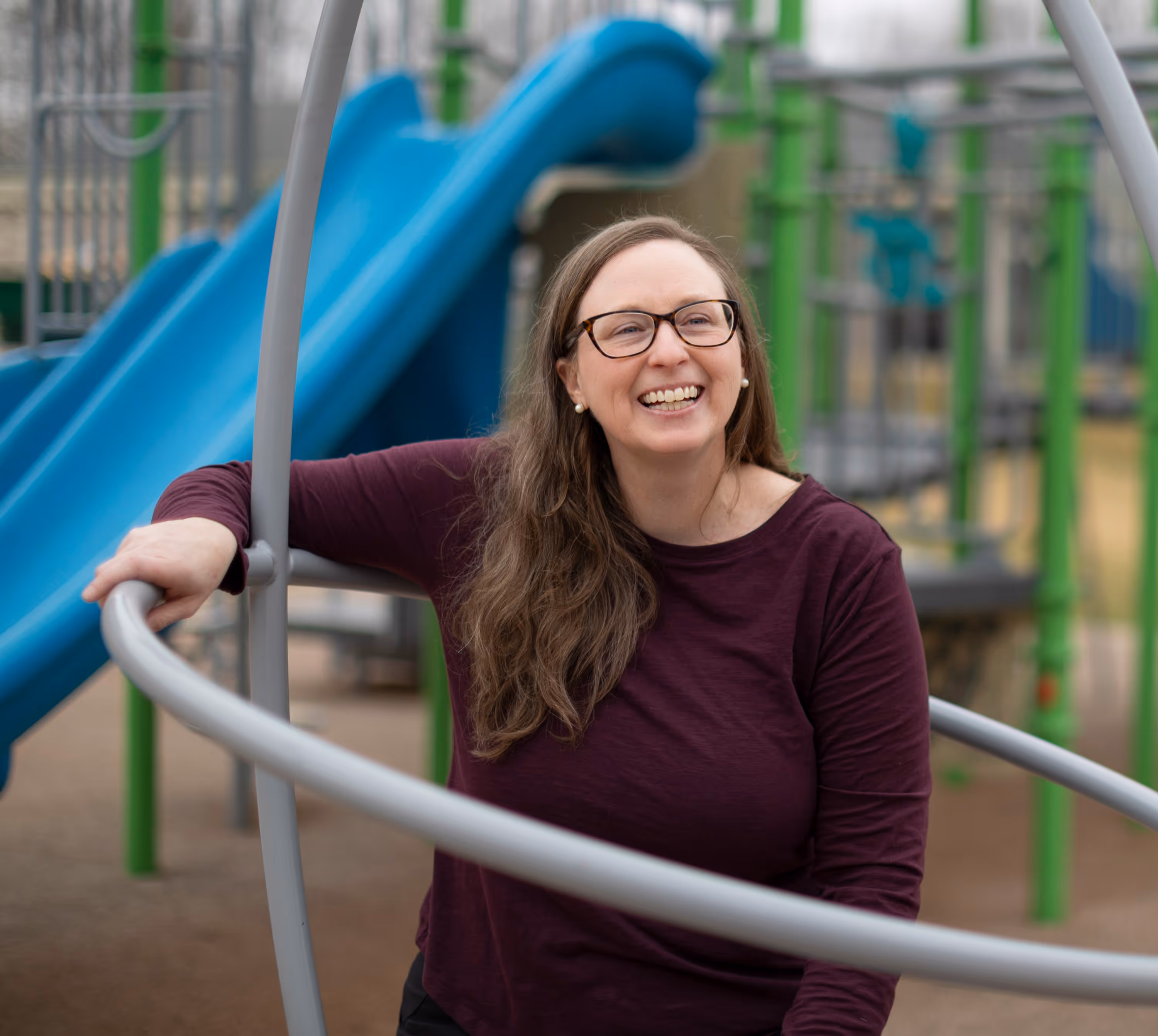 Smiling woman in glasses and maroon shirt at playground near blue slide and green poles.