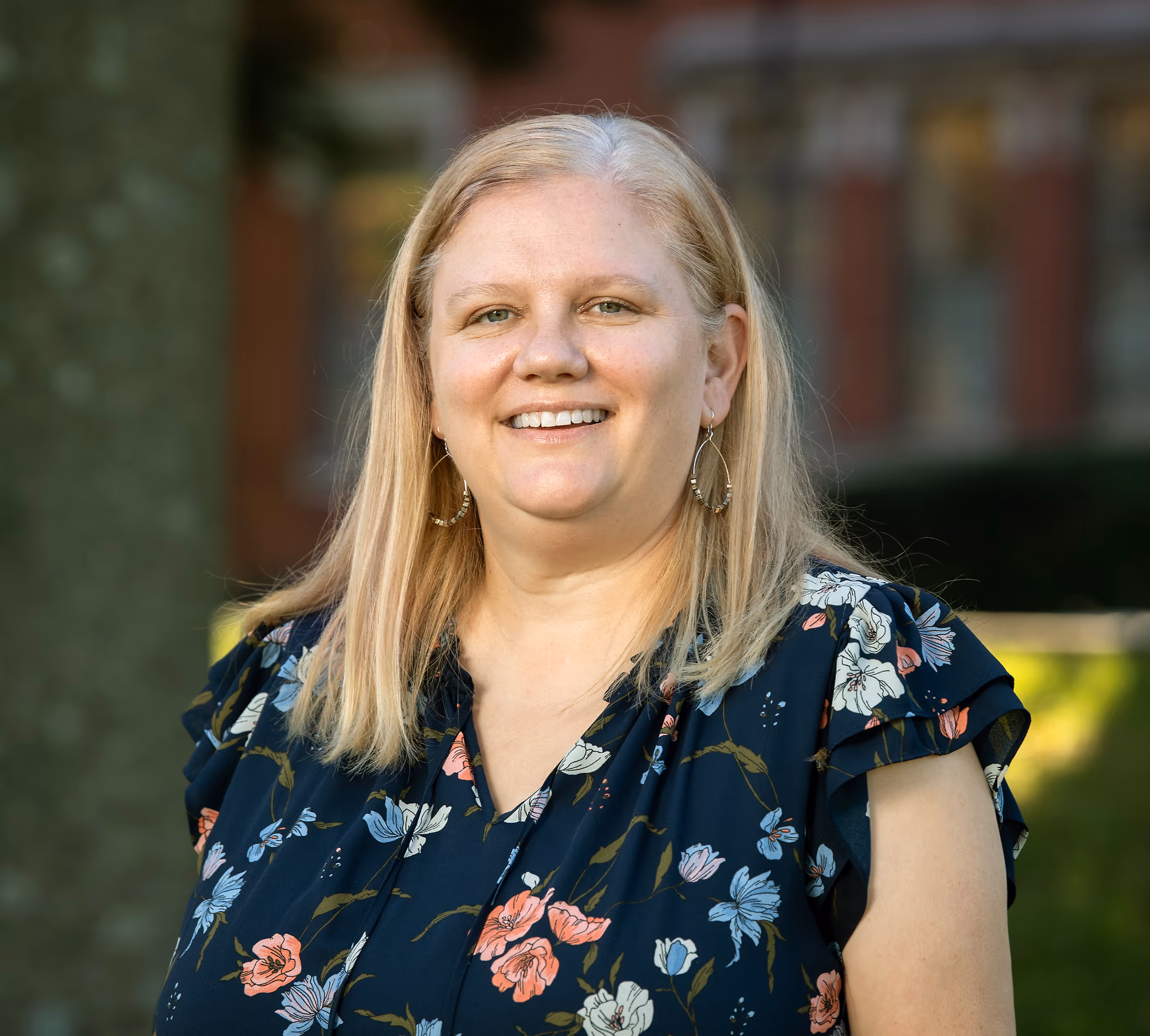 Smiling woman with shoulder-length blonde hair wearing floral blouse and hoop earrings outdoors.