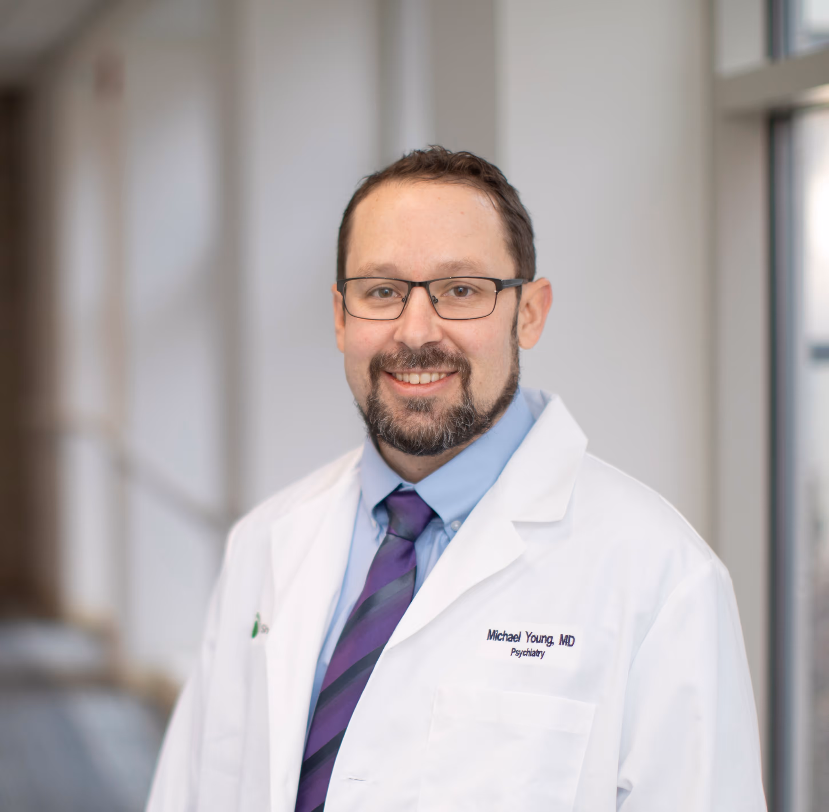 Smiling male doctor wearing glasses and white coat standing in a bright hospital corridor.