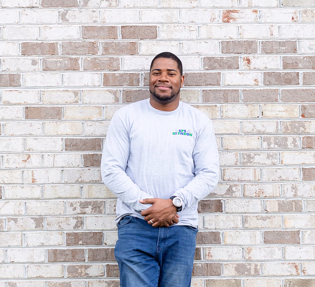 Man with short hair and beard in white long sleeve and jeans standing against brick wall.