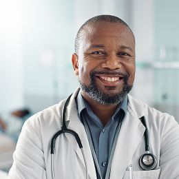 Smiling male doctor wearing a white coat and stethoscope in a bright medical office.