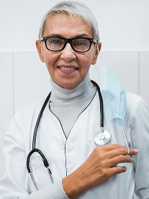 Smiling female doctor with short gray hair, glasses, stethoscope around neck, and holding a face mask.