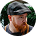 Bearded man wearing glasses, a black cap, and an orange shirt, looking to the side against a green background.