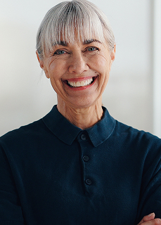 Smiling older woman with short gray hair wearing a dark polo shirt against a light background.