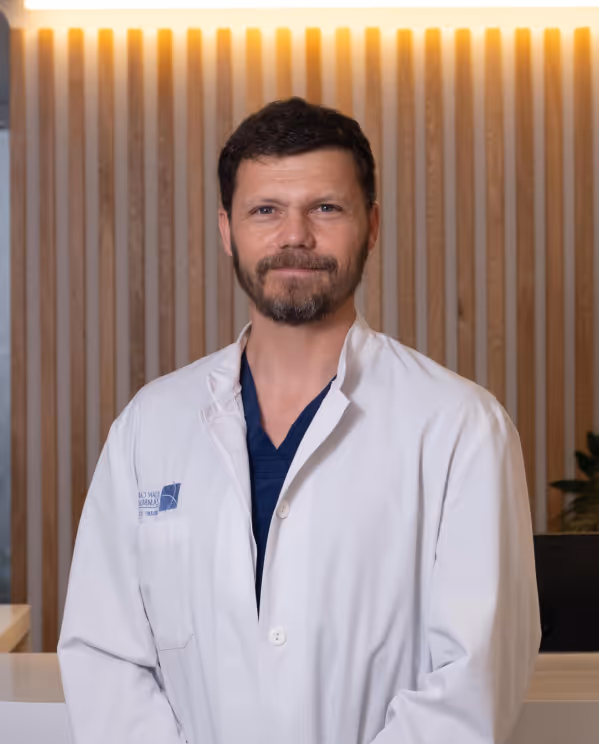 Man with short dark hair and beard wearing a white medical coat over a navy blue shirt, standing indoors with a wood-paneled background.