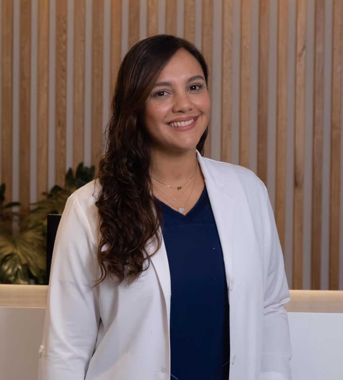 Smiling woman with long wavy hair wearing a white coat and navy blue top standing in front of a wooden slat wall.