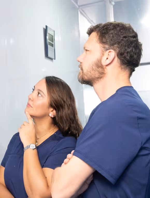 Two medical professionals in blue scrubs looking thoughtfully at a wall-mounted monitor.