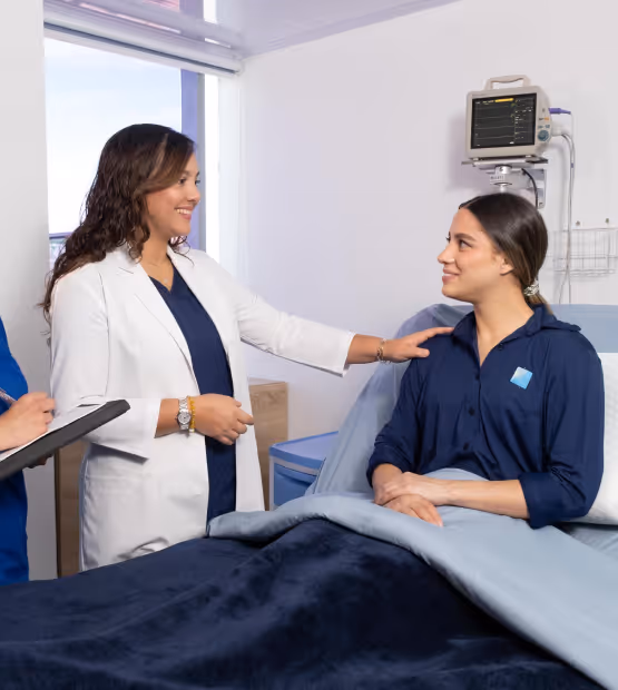 Healthcare professional in white coat smiling and reassuring a female patient sitting in a hospital bed covered with a blanket.
