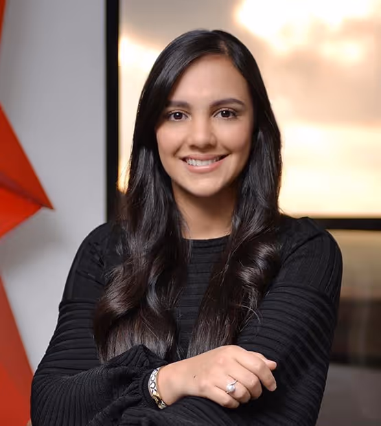Smiling woman with long dark hair wearing a black top, posing indoors with arms crossed.