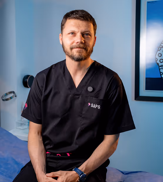 Bearded man wearing black medical scrubs with SAPS logo sitting on a medical examination bed.