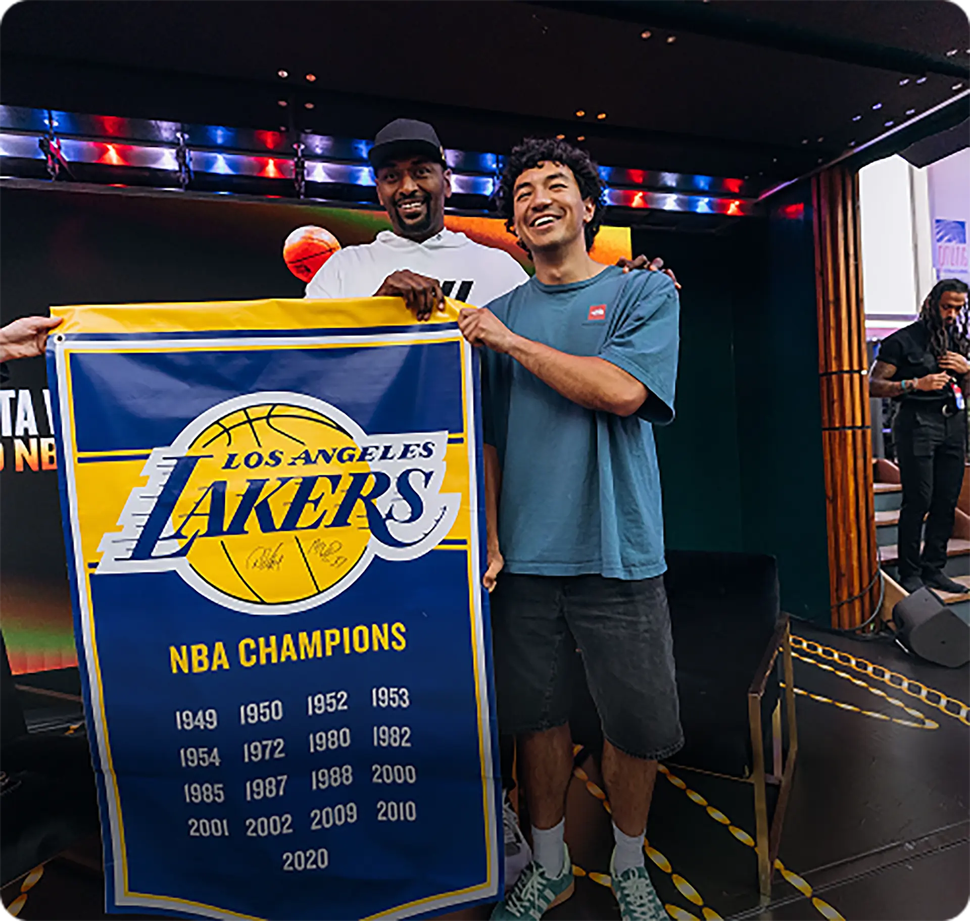 Two men smiling and holding a Los Angeles Lakers NBA Champions banner with championship years listed.