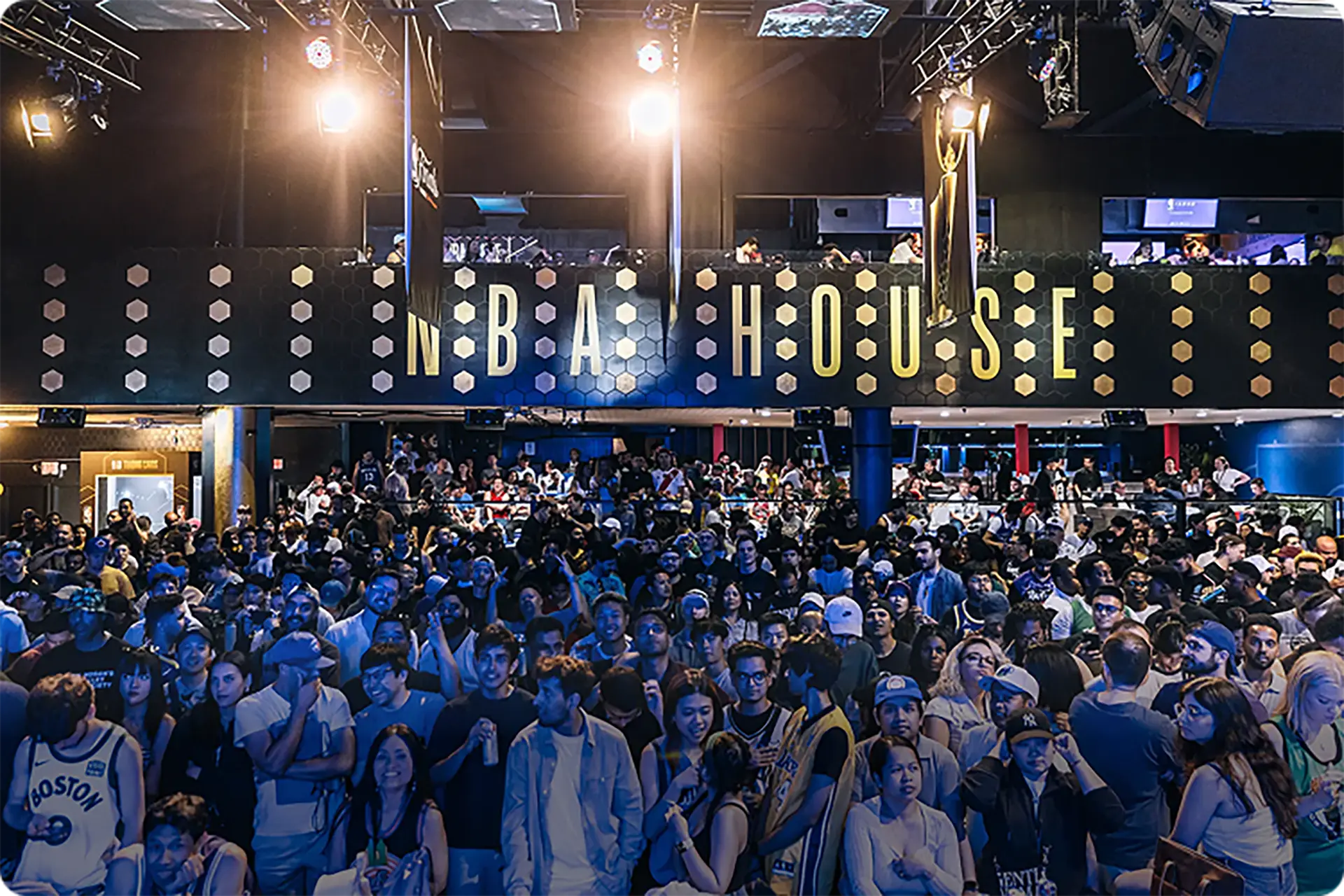 Crowd of diverse people gathered in a dimly lit venue with large 'NBA HOUSE' sign above.