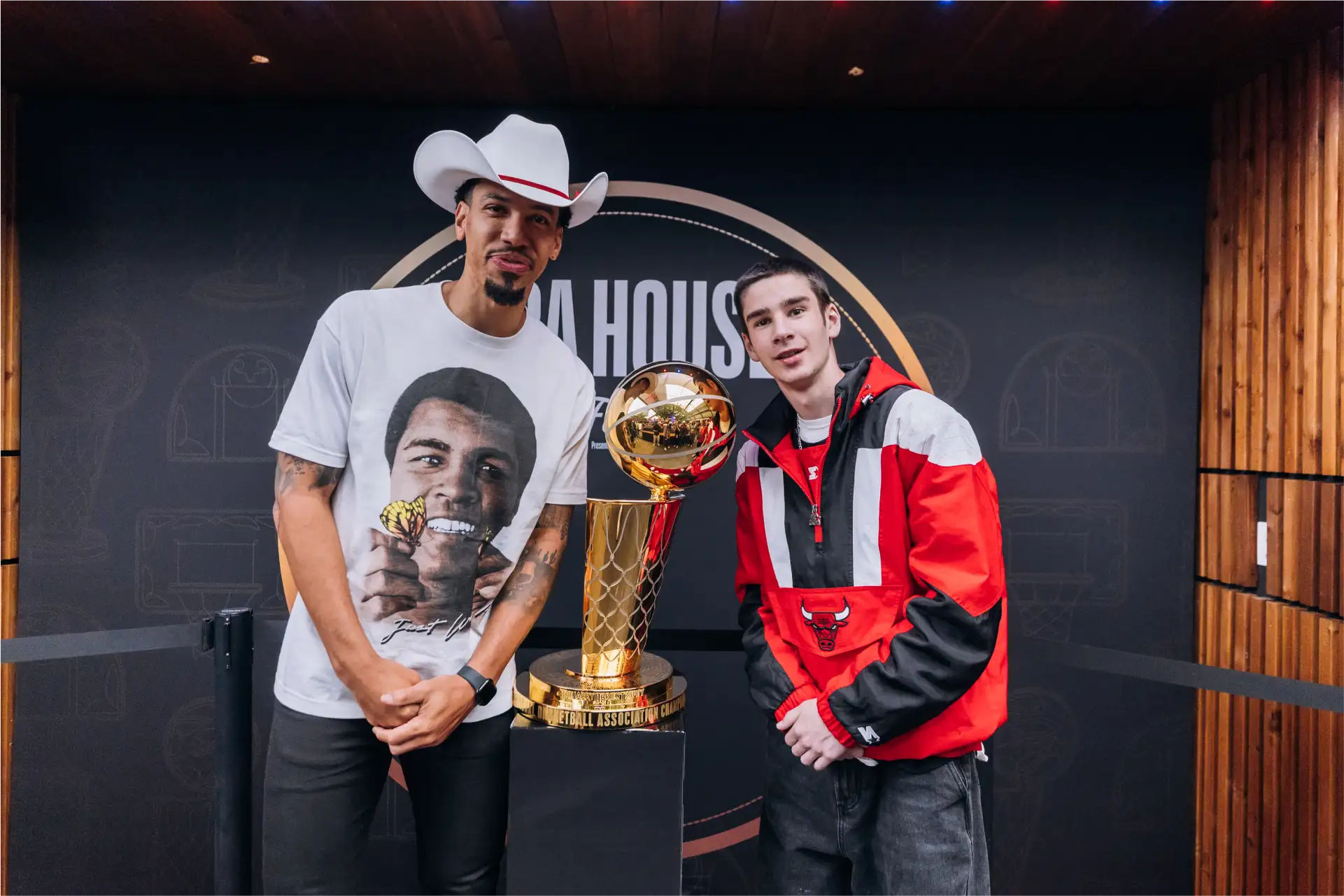 Two young men standing on either side of the Larry O'Brien NBA Championship Trophy; one wears a white cowboy hat and a t-shirt with a face and butterfly graphic, and the other wears a red Chicago Bulls jacket.