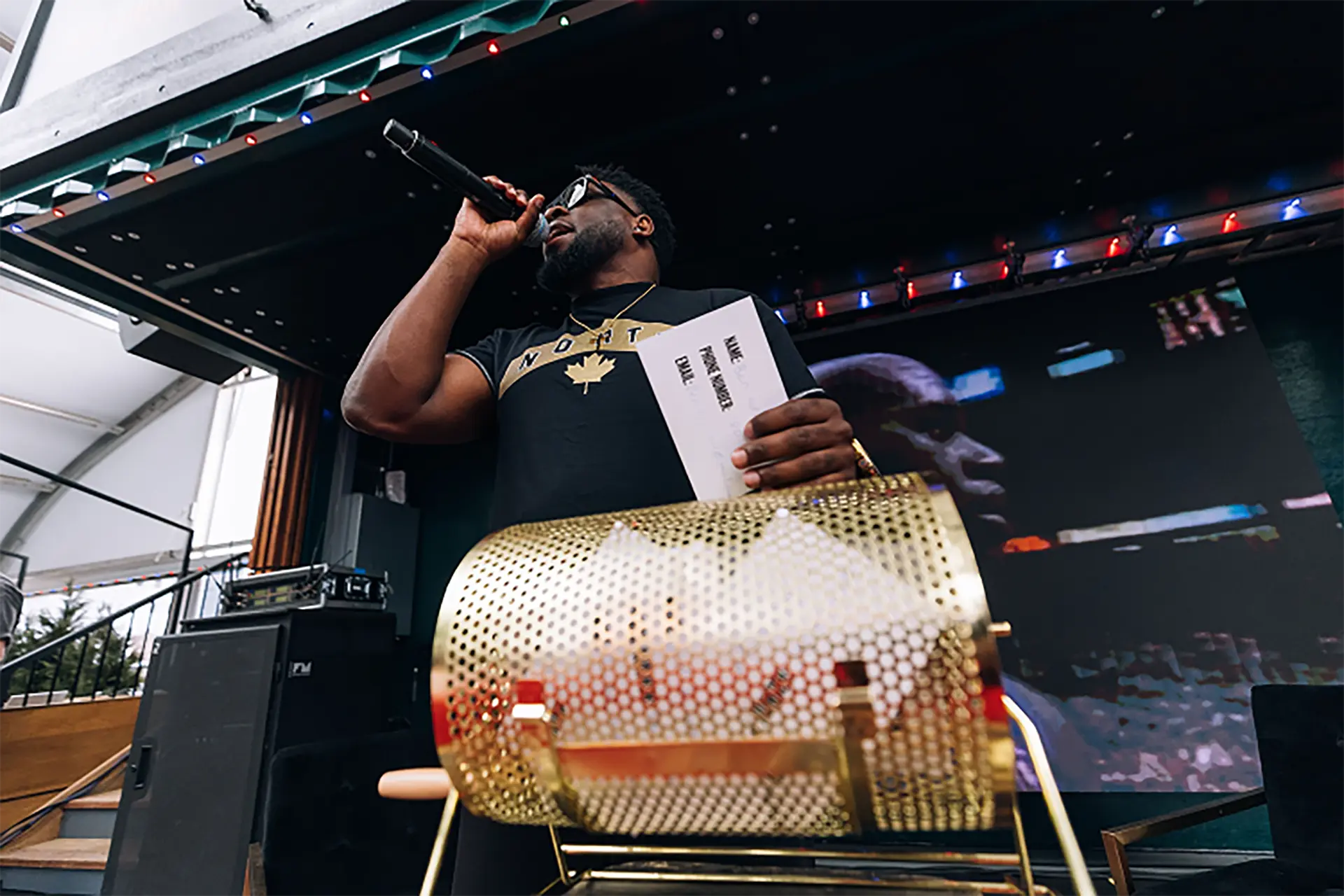 Man speaking into a microphone while holding a ticket near a gold raffle drum on a stage.