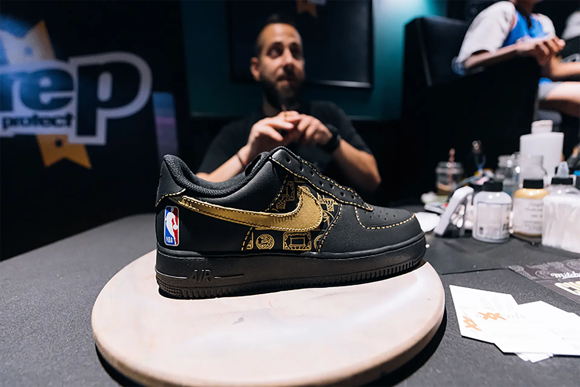 Black and gold Nike sneaker with NBA logo on heel displayed on a round wooden platform in a workshop.