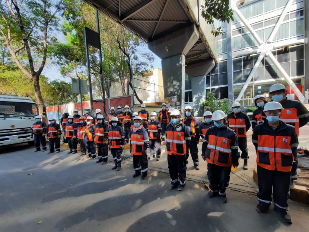 Group of construction workers wearing white helmets, face masks, and orange safety vests standing in a line under a bridge near a building and trees.