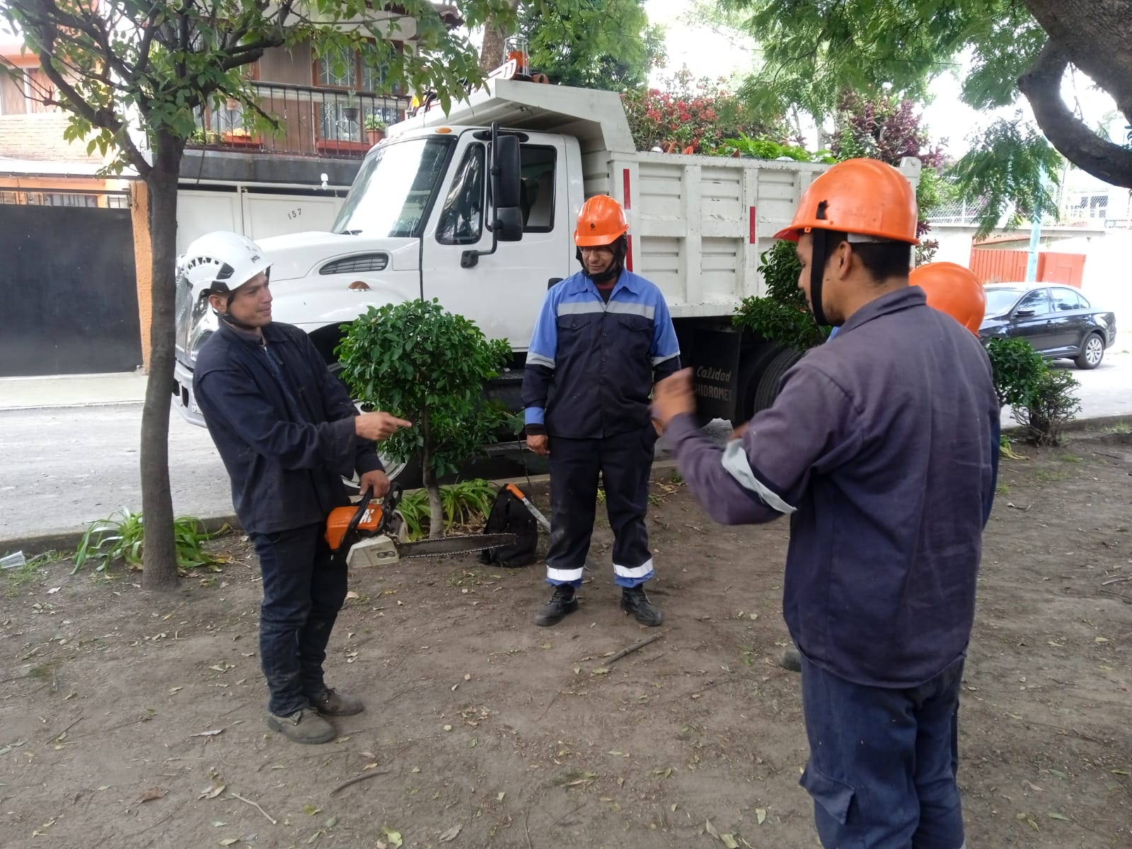 Four workers wearing helmets and uniforms standing outdoors near a white dump truck with one holding a chainsaw.