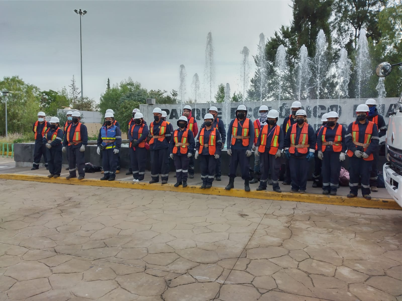 Group of workers wearing safety helmets, orange reflective vests, masks, and gloves standing in a line outdoors near a fountain.
