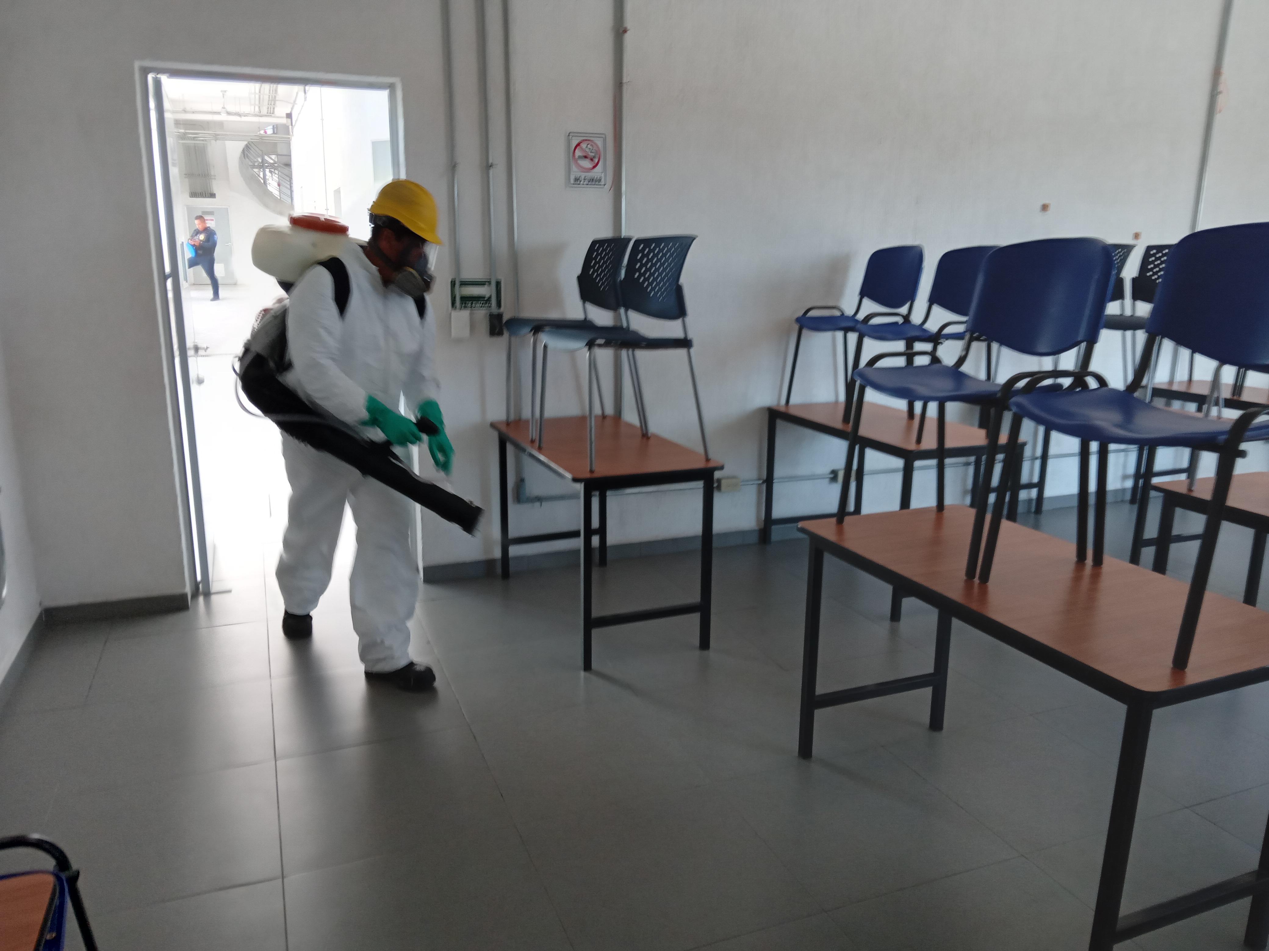 Person in protective suit, mask, gloves, and helmet disinfecting a room with blue chairs on tables.
