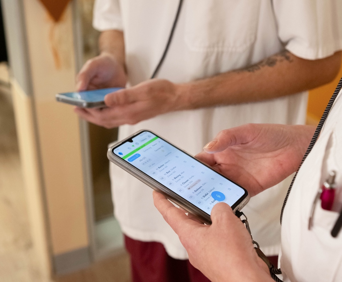 Hände einer Pflegekraft halten ein Smartphone mit der voize-App; im Hintergrund eine zweite Person mit Telefon.