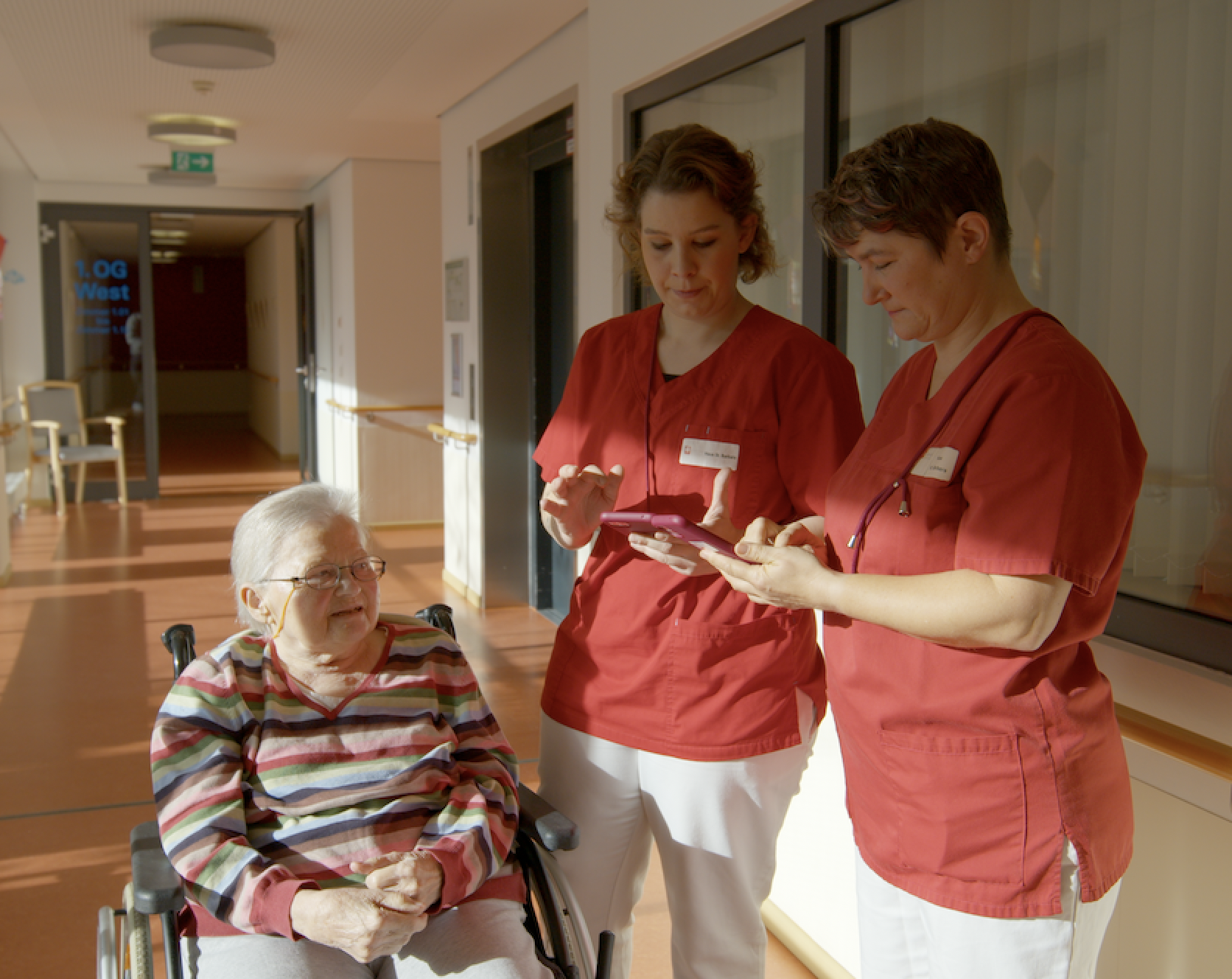 Drei Pflegekräfte in roter und weißer Dienstkleidung gehen einen Flur entlang, eine mit Smartphone.