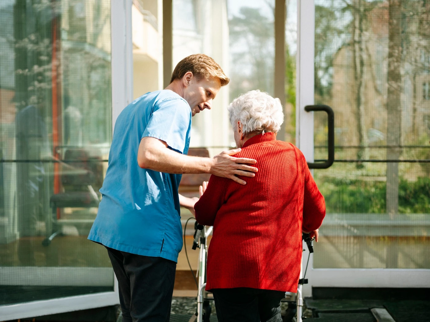 Pflegekraft hilft älterer Frau mit Rollator beim Gehen vor einem Glastür-Eingang.