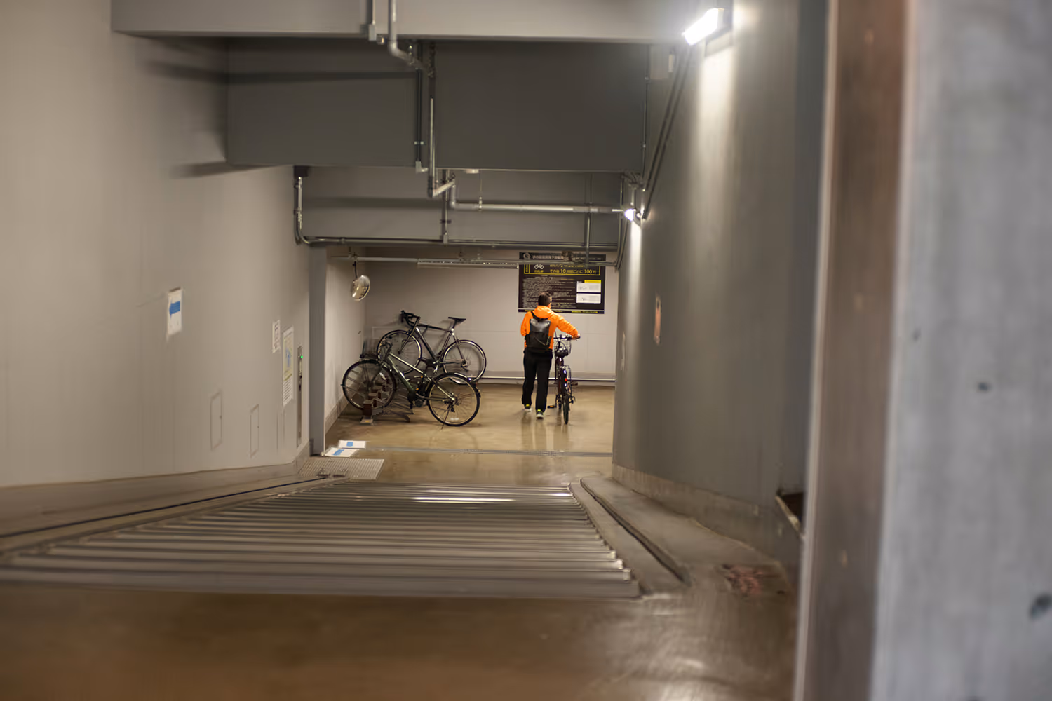 Person in an orange jacket standing with a bicycle in a dimly lit indoor bike storage area with several parked bikes.